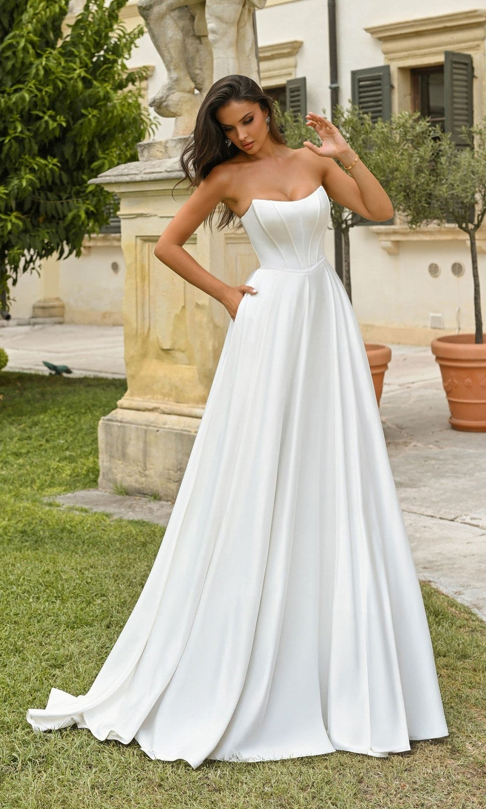 A woman stands outdoors near a stone statue and potted plants wearing the Alyce 7110 long dress in Diamond White, featuring a fitted bodice, full satin A-line skirt, and elegant strapless formal design.
